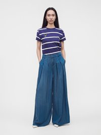 Femme debout, mains dans les poches, portant un t-shirt rayé bleu marine et blanc et un pantalon bleu à jambes larges sur un fond uni.