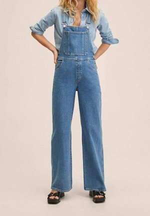 Frau trägt hellblaue Jeans-Latzhose und ein hochgekrempeltes langärmeliges Hemd, steht mit den Händen in der Hüfte vor einem einfarbigen Hintergrund.