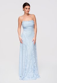 Femme debout portant une robe longue jusqu'au sol, bleu clair, sans manches, avec de fines bretelles et des motifs floraux subtils sur un fond uni.