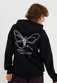 Felpa nera con cappuccio caratterizzata da un design di una farfalla bianca sul retro, con la scritta "NON FINIRE MAI NIENTE" sotto. Materiale morbido e vestibilità rilassata.