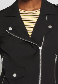 Zwarte motorjack met zilveren ritsen en drukknopen. Bevat een hoge kraag, diagonale ritssluiting en een vo pocket. Gestreepte top eronder.