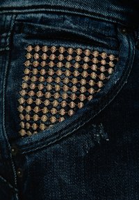 Mörkblå denimjeans har en ficka prydd med ett rutmönster av små, blanka bronsnitar som förbättrar textur och visuell intresse.