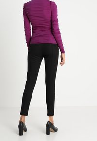 Eng anliegendes, langärmliges Oberteil in Lila mit geraffter Textur; kombiniert mit schwarzen, schmal geschnittenen Hosen und High Heels, die eine schlanke Silhouette betonen.