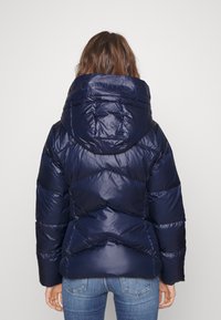 Marinblå puffjacka med huva, med en glansig yta, kviltat mönster och chevron-design, som visar detaljerad sömnad på ryggen.