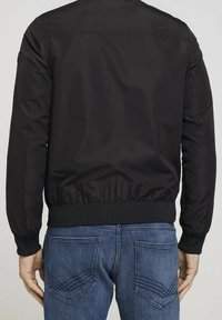 Černá bomber bunda s hladkým povrchem, žebrovanými manžetami a pasem. Má zadní díl yoke a žádné viditelné značky ani kovové doplňky.