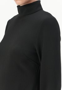 Haut noir à col montant en tissu lisse ; présente des détails de couture et une poche latérale subtile. Coupe cintrée avec des manches longues.
