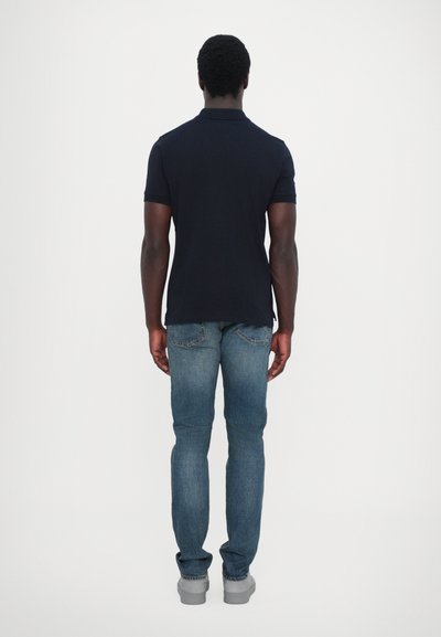 Navy polo shirt with short sleeves, paired with light blue jeans. The outfit is completed with gray shoes, viewed from the back.
