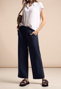 Kvinna som bär en vit kortärmad skjorta med knappar, marinblå vida byxor, svarta sandaler med spänne och bär en brun vävd axelremsväska.