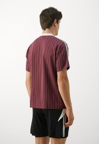T-shirt de sport rayé bordeaux à manches courtes, avec des accents blancs sur les épaules, coupe décontractée et texture lisse. Il possède un col ras du cou.