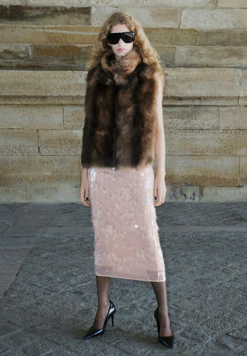 Gilet en fausse fourrure marron, porté avec une jupe midi en sequins rose et des escarpins noirs à talons aiguilles. La mannequin porte de grandes lunettes de soleil noires.
