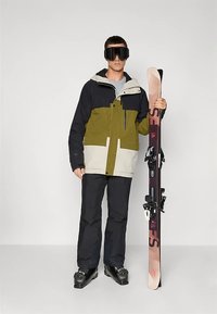 Man in een zwart en groen ski-jack en -broek, die een paar ski's vasthoudt, met zwarte skiboots en grote zwarte skibril.