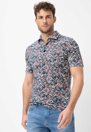 Homme portant un polo à manches courtes avec col, à motif floral bleu, rose et blanc, associé à un jean bleu.