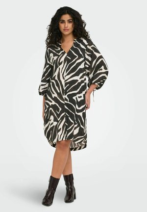 Femme debout aux cheveux bouclés portant une robe mi-longue imprimé zèbre noir et blanc et des bottines noires sur un fond uni.