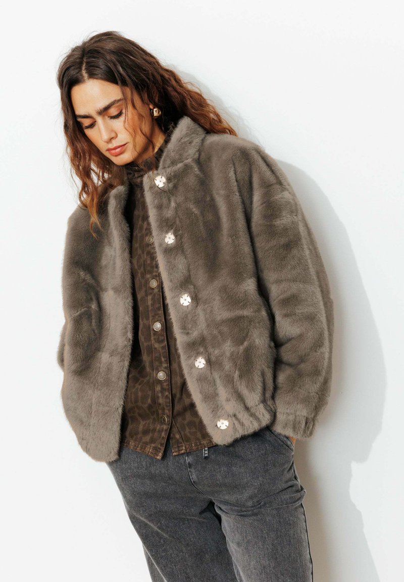 Veste en fausse fourrure grise avec un col haut, dotée de fermetures à boutons et d'une texture pelucheuse, superposée à une chemise à motifs sombres.