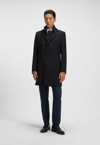 Cappotto in lana nera con rever a toppa, design a un solo petto e tre bottoni. Indossato sopra un completo, con pantaloni scuri e scarpe.