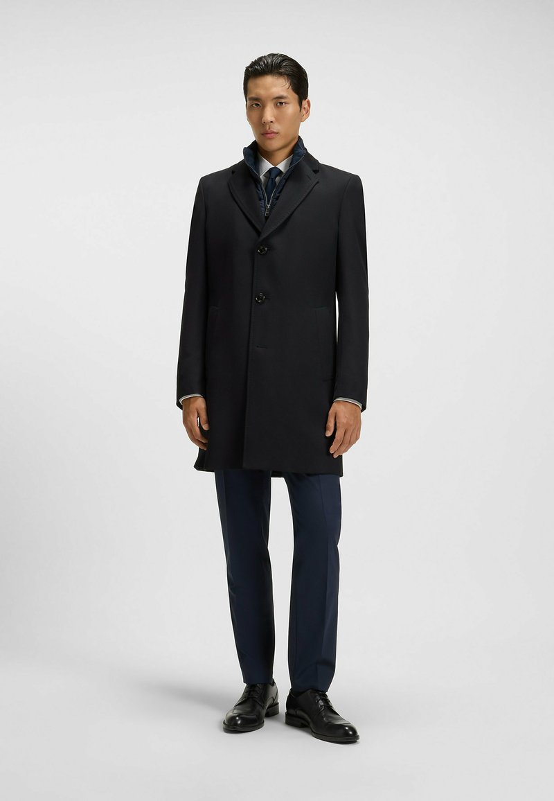 Cappotto in lana nera con rever a toppa, design a un solo petto e tre bottoni. Indossato sopra un completo, con pantaloni scuri e scarpe.