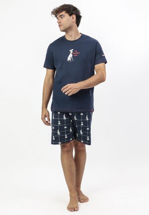 Hombre vistiendo una camiseta azul marino con un gráfico de perro y el texto "101 razones por las que" y pantalones cortos de cuadros azul marino con estampado de perros a juego, descalzo, de pie sobre fondo blanco.