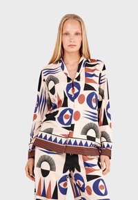 Patterned loungewear set featuring geometric designs in red, blue, and black on a beige background. Button-up top with a collar and striped cuffs.
