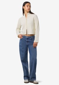 Veste cropped côtelée crème avec un col montant et une fermeture éclair devant, associée à un jean droit bleu clair et des baskets blanches.