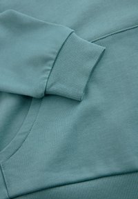 Türkizkék pulóver bordázott mandzsettával, sima textúrával és nagy kenguru zsebbel, laza szabású kialakítással.