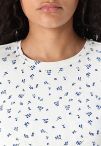 Vit ribbad topp med små blå blommönster, rund halsringning och subtil textur. Lämplig för vardagsbruk.