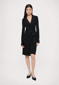 Čierne zavinovacie šaty s dlhými rukávmi, hlbokým výstrihom do V a opaskom v páse. Materiál pôsobí hladko; má elegantný strih a rozšírený lem.