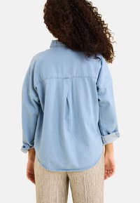 Camicia di jeans blu chiaro con maniche lunghe arrotolate e orlo curvato. Presenta uno spacco posteriore e una vestibilità rilassata. Indossata con pantaloni beige a righe.