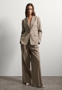 Completo beige su misura con blazer a un bottone, rever a lancia e pantaloni a gamba larga. Tessuto liscio, design minimalista e due tasche.
