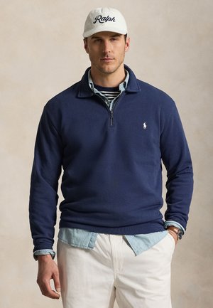 Mand iført hvid Ralph-kasket, marineblå kvart-lynlåssweater over lyseblå skjorte og stribet t-shirt, hvide bukser, med den ene hånd i lommen.