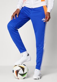 Blue athletic pants with white stripes down the sides, made of smooth fabric. A logo features on the thigh. White soccer cleats on a multicolored ball.