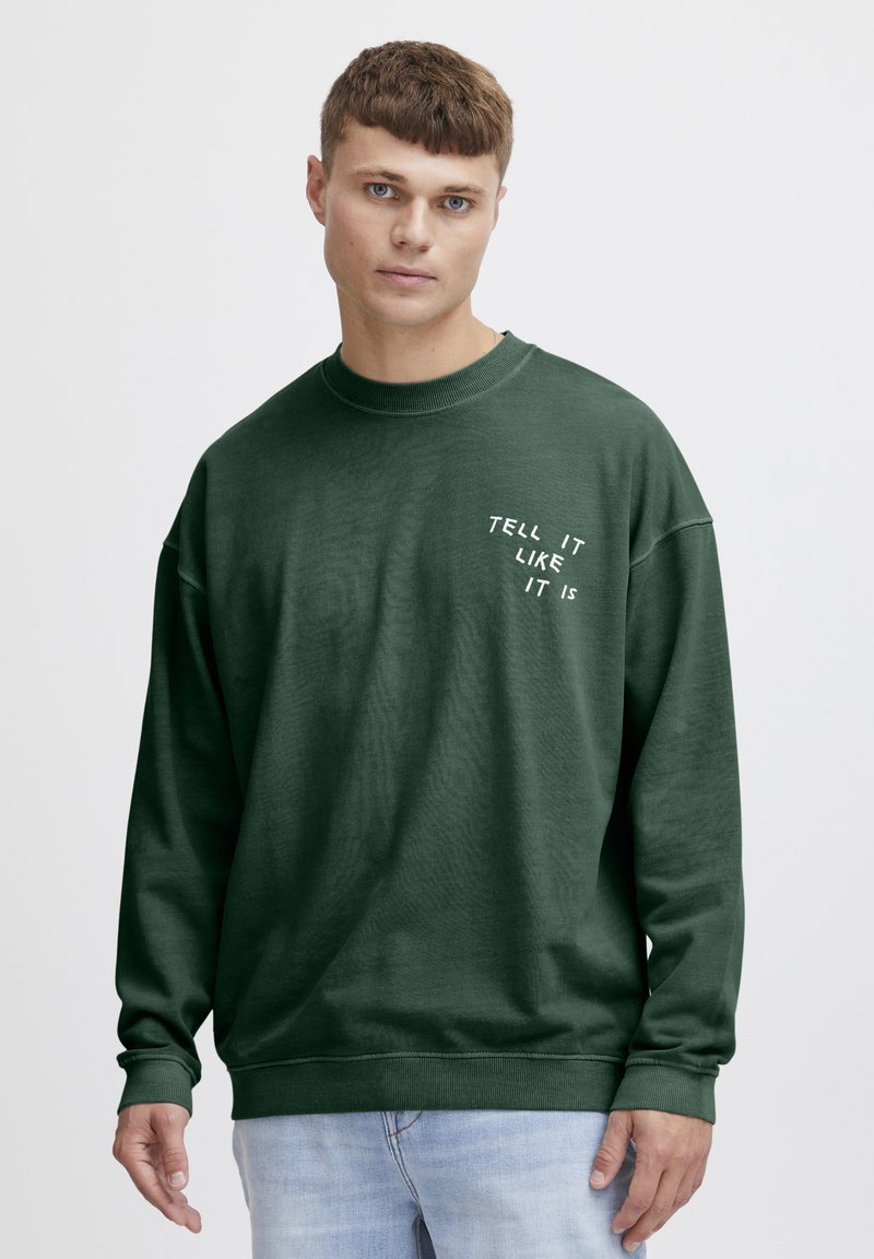 Sudadera verde con cuello redondo y mangas largas, que presenta el texto blanco "DILO COMO ES" en el pecho, hecha de una tela suave.