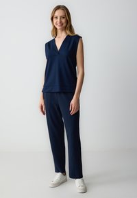 Top smanicato blu navy con scollatura a V e design a strati, abbinato a pantaloni dritti coordinati e sneakers bianche. Tessuto morbido.