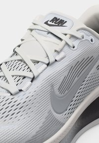 Λευκός αθλητικός παπούτσι Nike για τρέξιμο με σχέδιο από δίχτυ, ανοιχτό γκρι σήμα Swoosh, επίπεδα λευκά κορδόνια και ετικέτα με τη λέξη «RUN» στη γλώσσα.