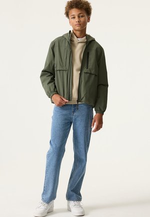 Olijfgroene, lichtgewicht jas met capuchon, voorzien van twee voorzakken en elastische manchetten. Gecombineerd met een beige hoodie en blauwe jeans.