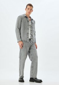 Chaqueta de mezclilla gris con cuello y cierre de botones, combinada con pantalones de mezclilla gris a juego y botas negras de suela gruesa.