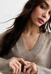 Beige v-hals trui met ribgebreide mouwen en onderzijde. Zacht, gestructureerd breimateriaal. Close-up van de halslijn en handen in een doordachte positie.