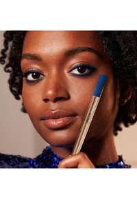 Niebieska kredka do oczu z metaliczną złotą obudową. Modelka ma ciemną skórę i kręcone włosy, a niebieska kredka podkreśla jej oczy.