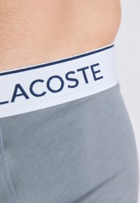Boxer-briefs en coton gris avec une ceinture élastique blanche sur laquelle est inscrit "LACOSTE" en lettrage marine sur le dessus. Texture lisse.