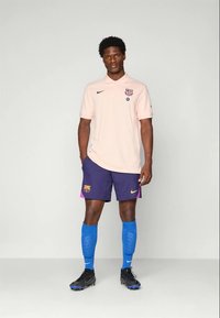 Man standing wearing light pink polo shirt, navy blue shorts with club logos, blue soccer socks, and black cleats against plain background.