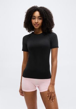 Jeune femme aux cheveux bouclés portant un haut noir ajusté à manches courtes et un short rose clair, debout devant un fond blanc uni.