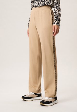 Beige wijde broek met een donkere chevron-zijkantstreep, gedragen met zwarte, beige en witte sneakers en een patroon lange mouwen top.