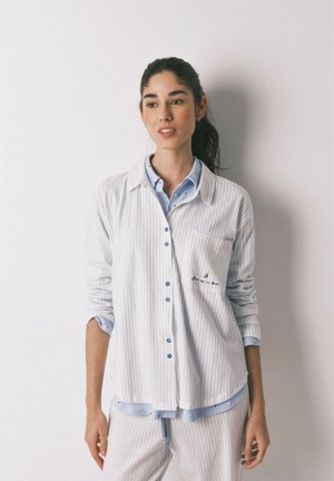 Kvinde med mørkt hår samlet i en hestehale, iført lyseblå trøje og hvid skjorte med tynde striber samt matchende bukser, stående foran en hvid væg.