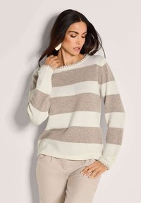 Frau in beigem und weißen horizontal gestreiften Pullover und beigen Hosen, die mit einer Hand nahe am Hals und der anderen auf der Hüfte posiert.