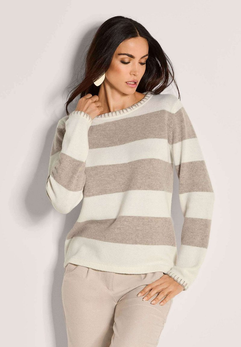 Frau in beigem und weißen horizontal gestreiften Pullover und beigen Hosen, die mit einer Hand nahe am Hals und der anderen auf der Hüfte posiert.