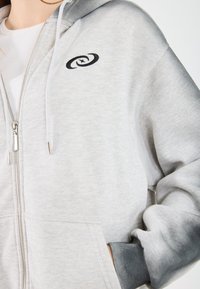 Felpa grigia con zip realizzata in tessuto testurizzato, dotata di una tasca frontale, coulisse regolabile e un logo nero. I dettagli includono polsini grigio scuro.