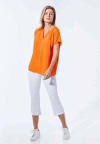 Oranje top met korte mouwen en V-hals, gecombineerd met witte cropped broeken en witte sneakers. Gladde stof zonder patronen of accenten.