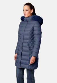 Abrigo de plumón azul marino con un diseño acolchado, con capucha forrada de piel, botones a presión y bolsillos laterales, combinado con jeans.