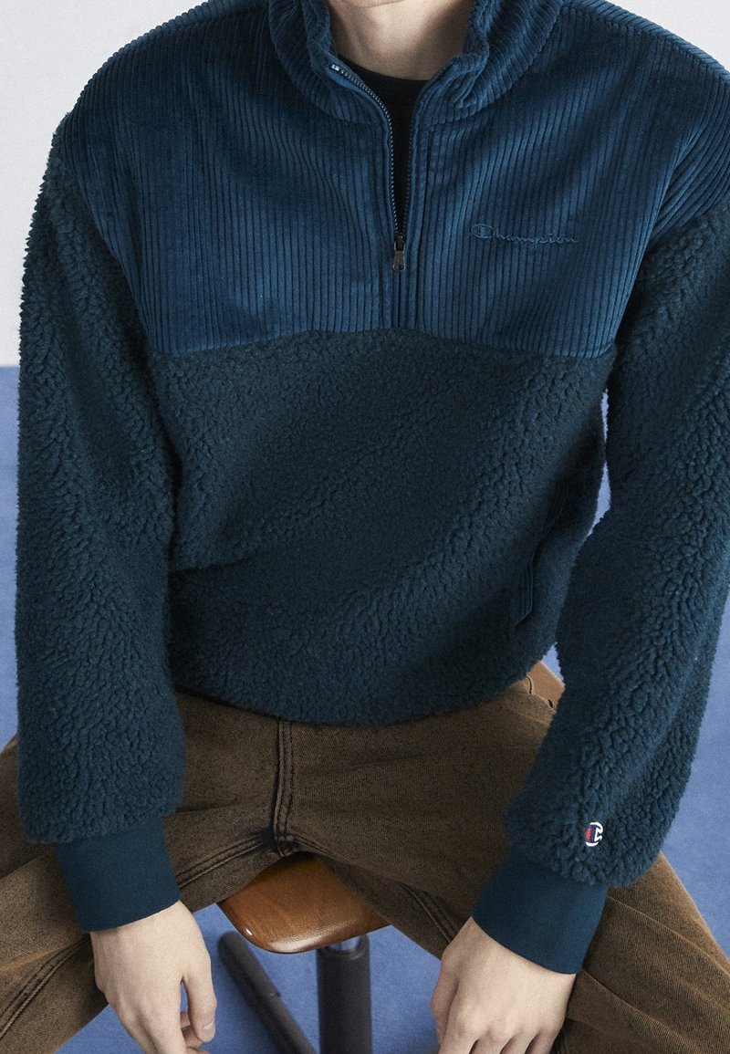 Mörkblå tröja med halv dragkedja, med ribbat övre parti och mjukt nedre, tillverkad av mjukt, strukturerat tyg. Litet logo på ärmen.