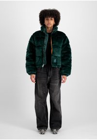 Jachetă puffer din blană sintetică verde, cu guler înalt, închidere cu fermoar și căptușeală portocalie în contrast. Purtați-o cu blugi negri cu picior larg și adidași.