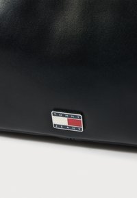 Μαύρη δερμάτινη τσάντα χειρός με ορθογώνιο σχήμα. Μπροστινό λογότυπο σε χρωματική παλέτα μπλε, λευκού και κόκκινου με το κείμενο "TOMMY JEANS".